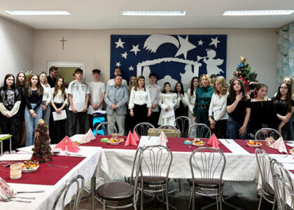 Grupa młodzieży i opiekunów stojących w sali przy świątecznie nakrytych stołach, podczas spotkania wigilijnego.