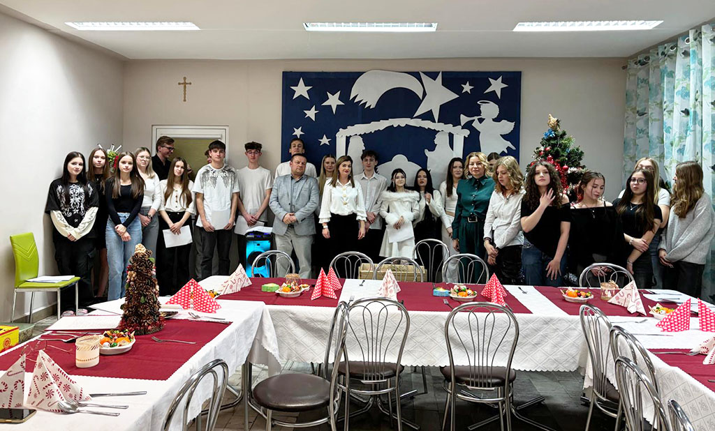 Grupa młodzieży i opiekunów stojących w sali przy świątecznie nakrytych stołach, podczas spotkania wigilijnego.