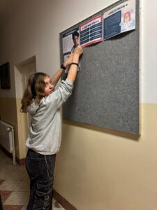 Grupa wychowanków bursy szkolnej prezentuje plakaty informacyjne dotyczące depresji oraz hasło „Stop depresji”. Na kolejnych zdjęciach jedna z uczestniczek przypina materiały edukacyjne na tablicy informacyjnej na korytarzu bursy. Plansze zawierają informacje o objawach depresji, sposobach szukania pomocy oraz profilaktyce.
