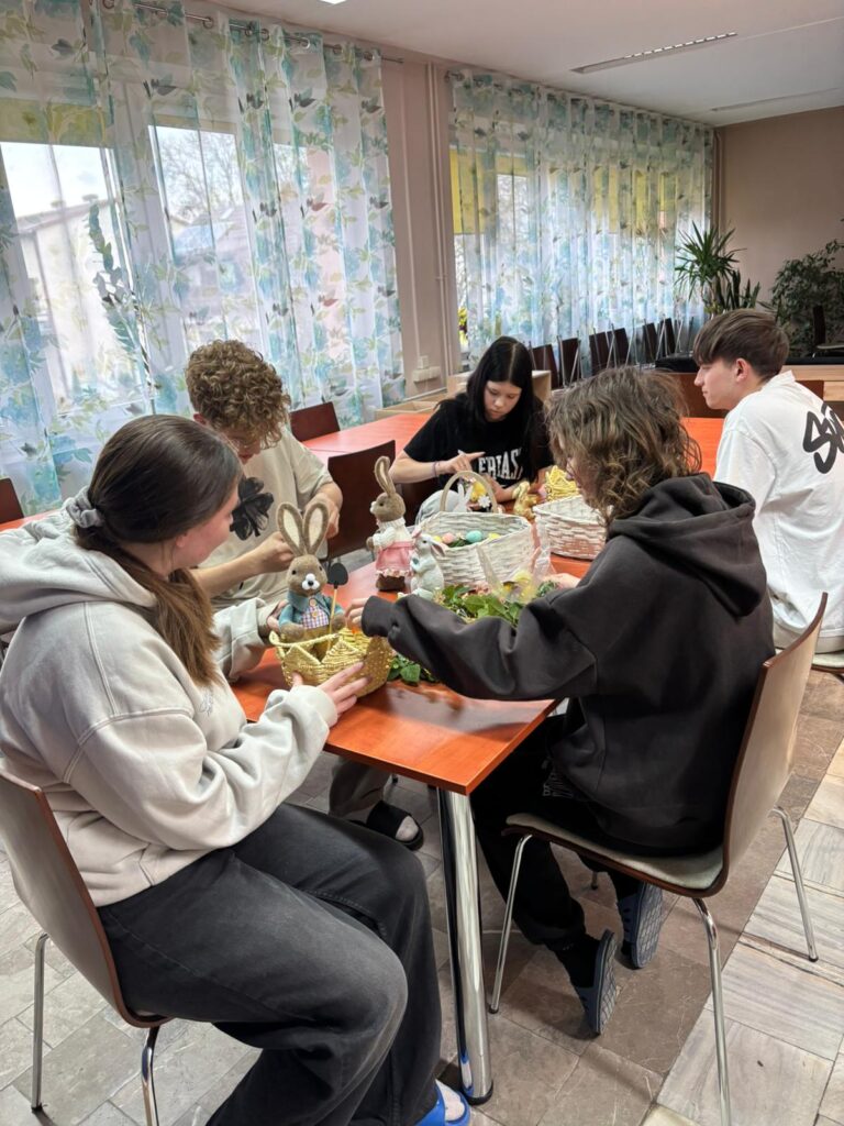 Wychowankowie bursy szkolnej podczas warsztatów wielkanocnych wykonują ręcznie stroiki świąteczne z motywami wiosennymi, takimi jak zajączki, pisanki i kurczaczki. Na zdjęciach widoczne są efekty ich pracy oraz wspólne zaangażowanie w tworzenie dekoracji w sali zajęć i przed budynkiem placówki