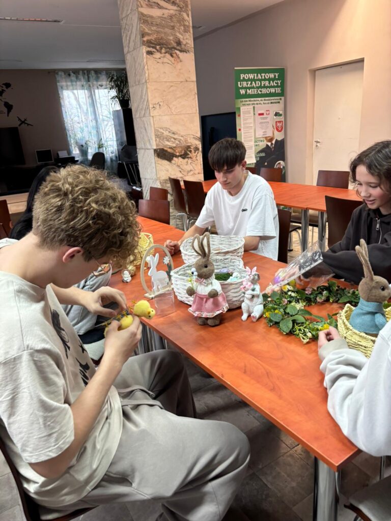 Wychowankowie bursy szkolnej podczas warsztatów wielkanocnych wykonują ręcznie stroiki świąteczne z motywami wiosennymi, takimi jak zajączki, pisanki i kurczaczki. Na zdjęciach widoczne są efekty ich pracy oraz wspólne zaangażowanie w tworzenie dekoracji w sali zajęć i przed budynkiem placówki