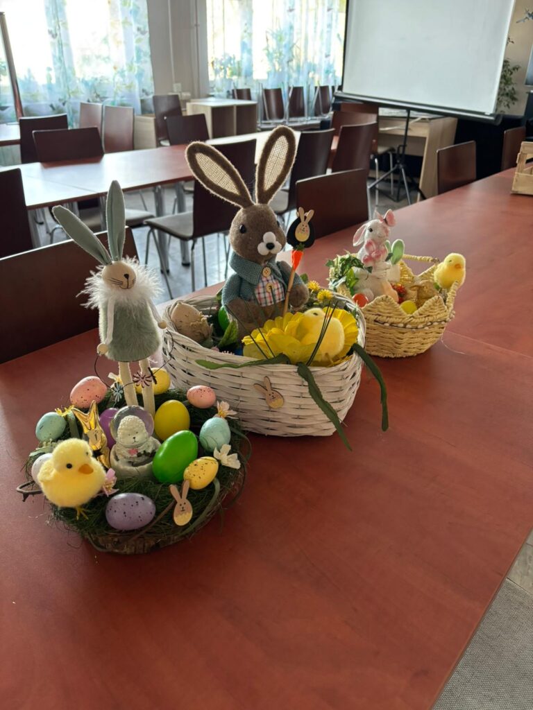 Wychowankowie bursy szkolnej podczas warsztatów wielkanocnych wykonują ręcznie stroiki świąteczne z motywami wiosennymi, takimi jak zajączki, pisanki i kurczaczki. Na zdjęciach widoczne są efekty ich pracy oraz wspólne zaangażowanie w tworzenie dekoracji w sali zajęć i przed budynkiem placówki