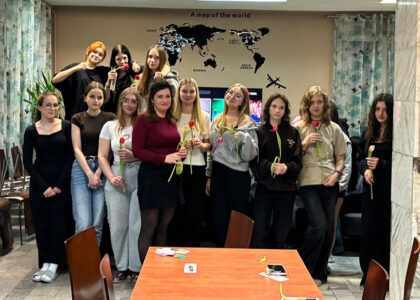 Grupa wychowanek bursy wraz z wychowawczynią podczas wspólnego zdjęcia z okazji Dnia Kobiet. Dziewczęta trzymają w dłoniach tulipany otrzymane z okazji święta i stoją w sali wspólnej bursy.