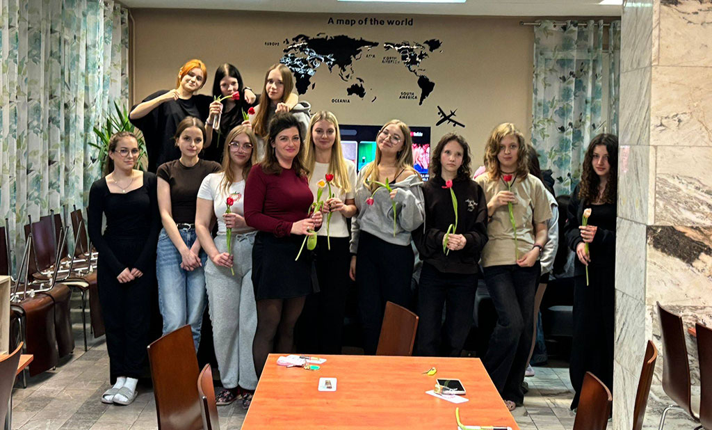 Grupa wychowanek bursy wraz z wychowawczynią podczas wspólnego zdjęcia z okazji Dnia Kobiet. Dziewczęta trzymają w dłoniach tulipany otrzymane z okazji święta i stoją w sali wspólnej bursy.