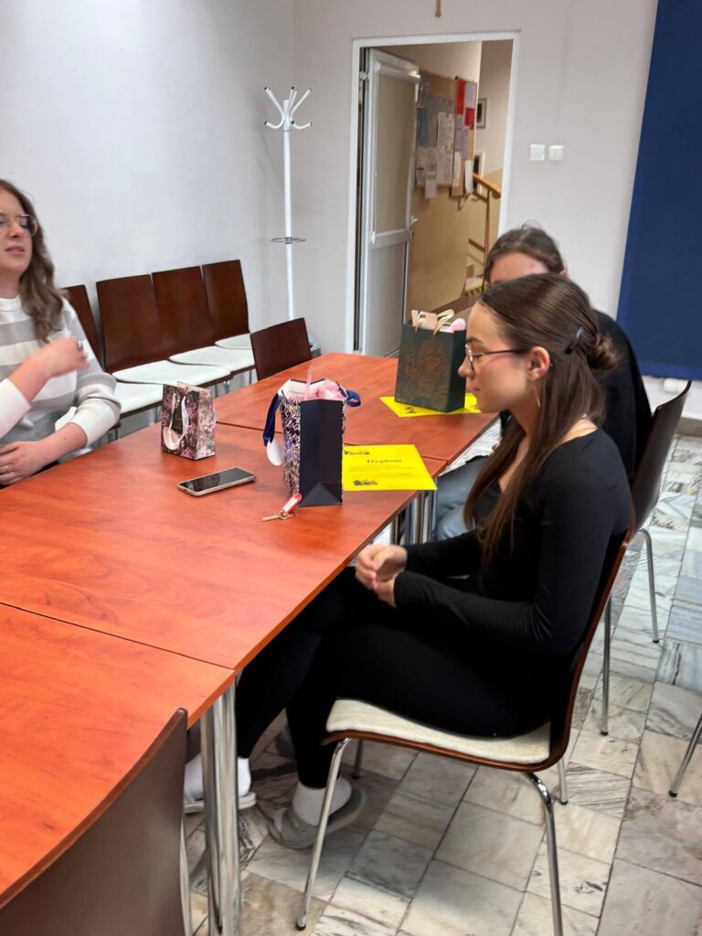 Zdjęcia przedstawiają uroczystość pożegnania maturzystów w Bursie Szkolnej. W sali z ustawionymi stołami i krzesłami grupa młodzieży oraz wychowawcy uczestniczą w oficjalnym spotkaniu. Na jednej z fotografii uczennica trzyma upominek, na innych widoczne jest wręczanie prezentów oraz odczytywanie przemówień i podziękowań. Na stołach znajdują się torby z upominkami. W tle widoczna jest tablica oraz elementy wyposażenia sali. Atmosfera wydarzenia ma charakter uroczysty i podniosły