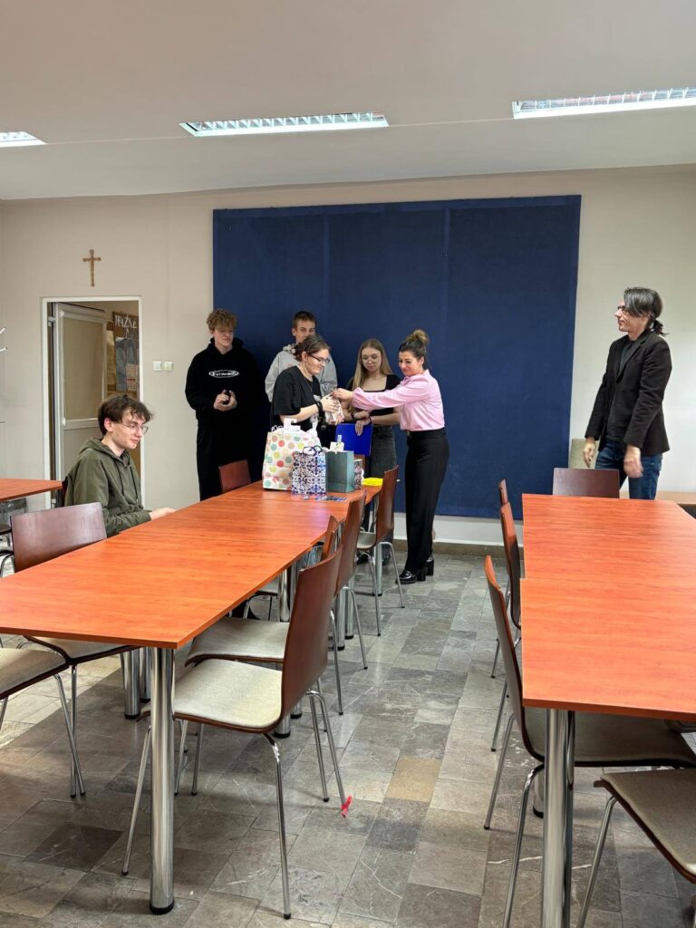 Zdjęcia przedstawiają uroczystość pożegnania maturzystów w Bursie Szkolnej. W sali z ustawionymi stołami i krzesłami grupa młodzieży oraz wychowawcy uczestniczą w oficjalnym spotkaniu. Na jednej z fotografii uczennica trzyma upominek, na innych widoczne jest wręczanie prezentów oraz odczytywanie przemówień i podziękowań. Na stołach znajdują się torby z upominkami. W tle widoczna jest tablica oraz elementy wyposażenia sali. Atmosfera wydarzenia ma charakter uroczysty i podniosły