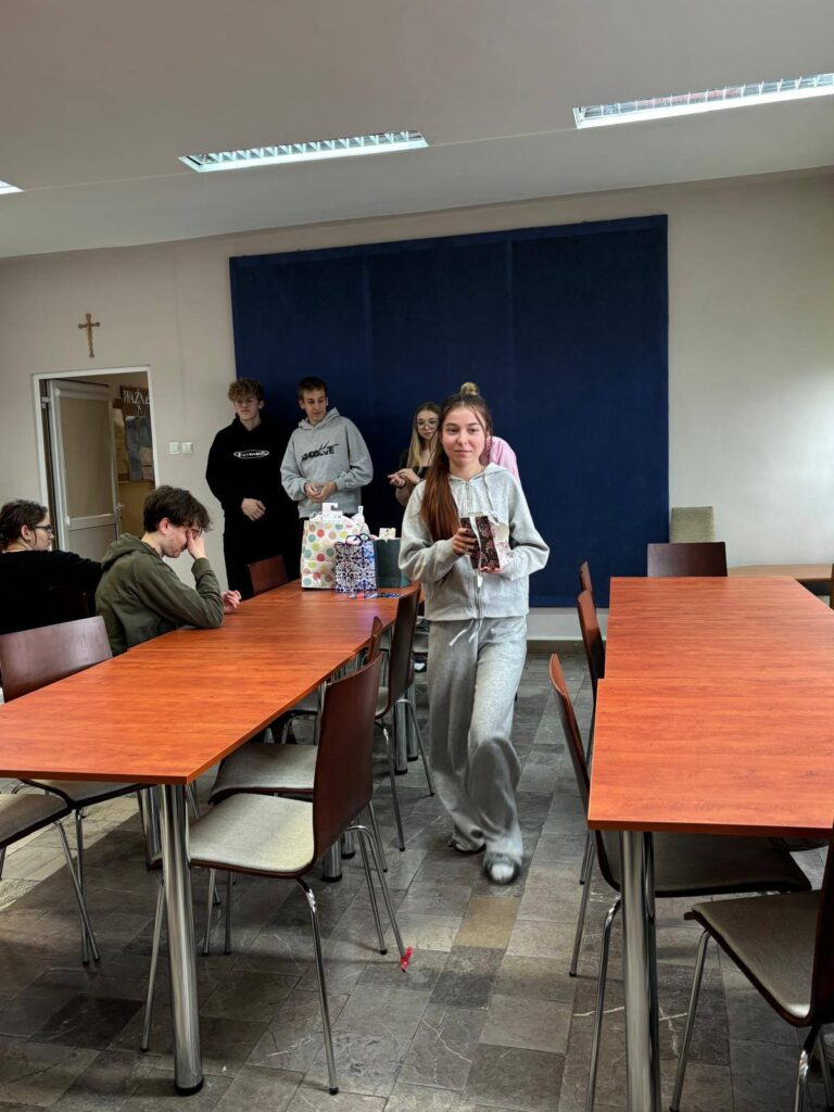 Zdjęcia przedstawiają uroczystość pożegnania maturzystów w Bursie Szkolnej. W sali z ustawionymi stołami i krzesłami grupa młodzieży oraz wychowawcy uczestniczą w oficjalnym spotkaniu. Na jednej z fotografii uczennica trzyma upominek, na innych widoczne jest wręczanie prezentów oraz odczytywanie przemówień i podziękowań. Na stołach znajdują się torby z upominkami. W tle widoczna jest tablica oraz elementy wyposażenia sali. Atmosfera wydarzenia ma charakter uroczysty i podniosły