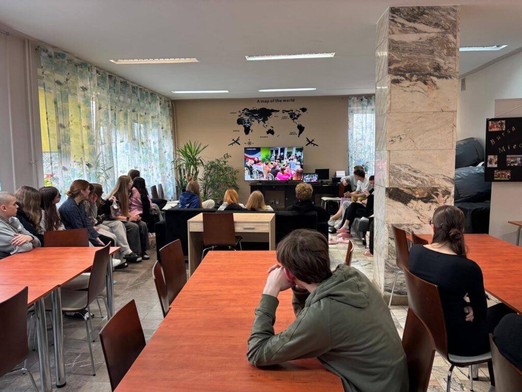 Zdjęcia przedstawiają uroczystość pożegnania maturzystów w Bursie Szkolnej. W sali z ustawionymi stołami i krzesłami grupa młodzieży oraz wychowawcy uczestniczą w oficjalnym spotkaniu. Na jednej z fotografii uczennica trzyma upominek, na innych widoczne jest wręczanie prezentów oraz odczytywanie przemówień i podziękowań. Na stołach znajdują się torby z upominkami. W tle widoczna jest tablica oraz elementy wyposażenia sali. Atmosfera wydarzenia ma charakter uroczysty i podniosły