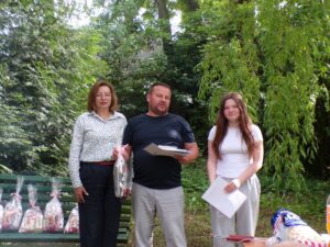 Zdjęcia z plenerowego spotkania integracyjnego w otoczeniu zieleni. Uczestnicy wspólnie spędzają czas na świeżym powietrzu, biorąc udział w części oficjalnej oraz przygotowując poczęstunek przy ognisku. W tle widoczne są drzewa i naturalne otoczenie sprzyjające rekreacji i integracji