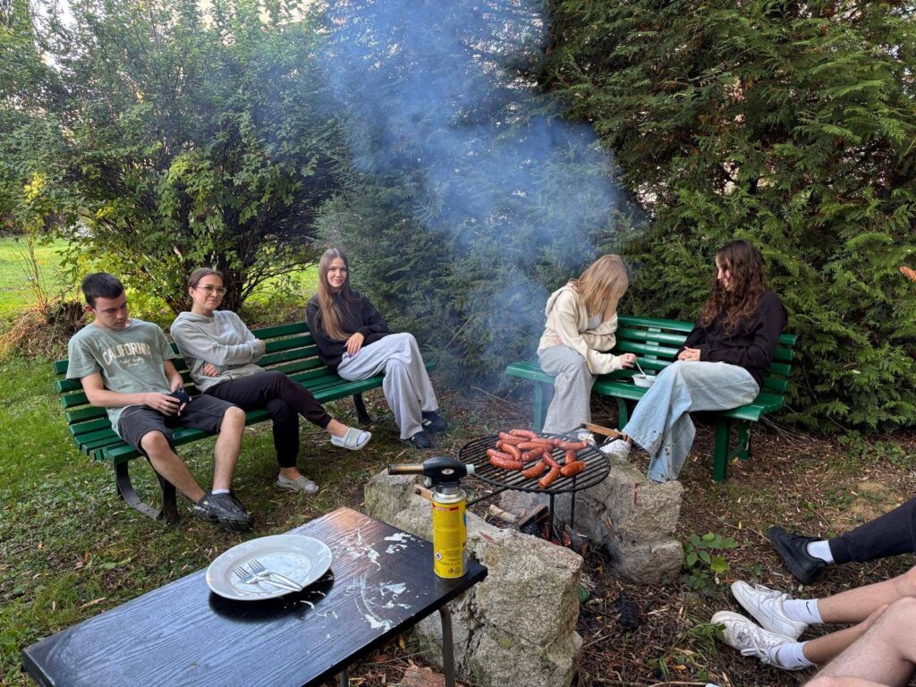 Grupa wychowanków Bursy Szkolnej podczas wspólnego ogniska z okazji Dnia Chłopaka. Młodzież siedzi na ławkach w otoczeniu zieleni, a na grillu nad ogniskiem pieką się kiełbaski.