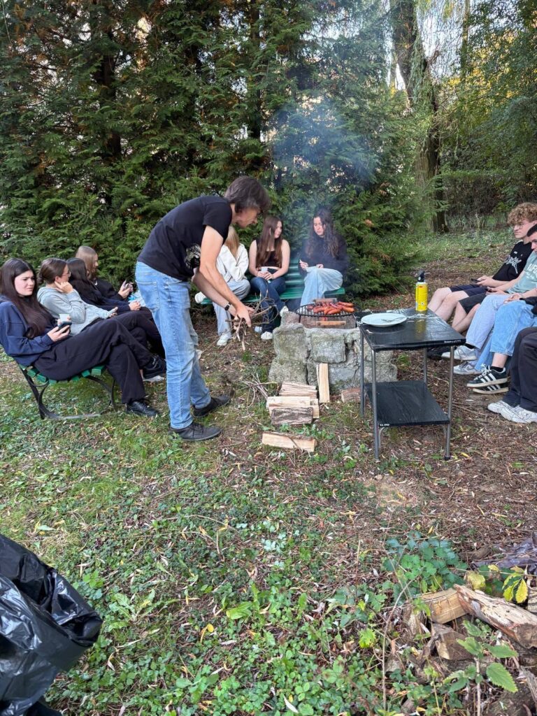 Grupa wychowanków Bursy Szkolnej podczas wspólnego ogniska z okazji Dnia Chłopaka. Młodzież siedzi na ławkach w otoczeniu zieleni, a na grillu nad ogniskiem pieką się kiełbaski.