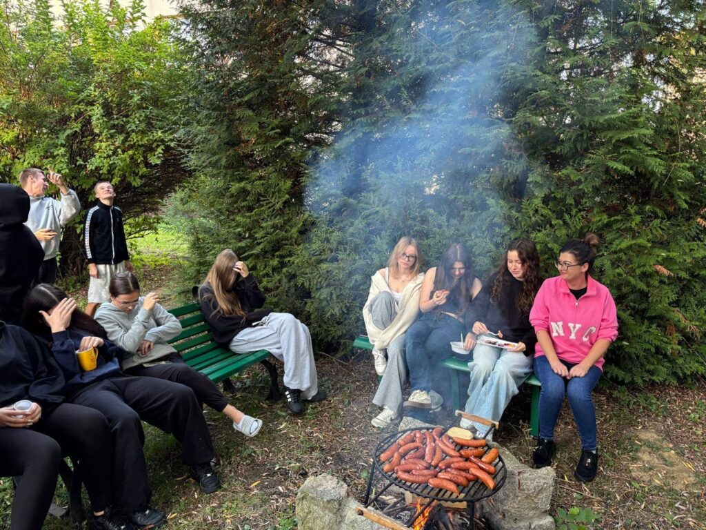 Grupa wychowanków Bursy Szkolnej podczas wspólnego ogniska z okazji Dnia Chłopaka. Młodzież siedzi na ławkach w otoczeniu zieleni, a na grillu nad ogniskiem pieką się kiełbaski.