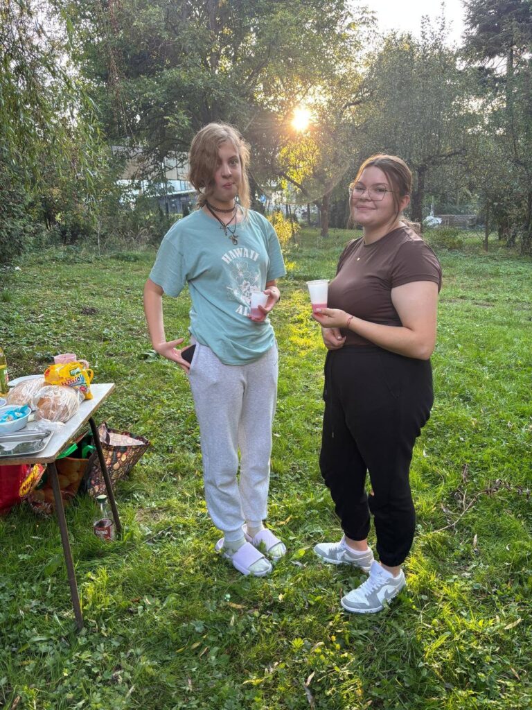 Grupa wychowanków Bursy Szkolnej podczas wspólnego ogniska z okazji Dnia Chłopaka. Młodzież siedzi na ławkach w otoczeniu zieleni, a na grillu nad ogniskiem pieką się kiełbaski.