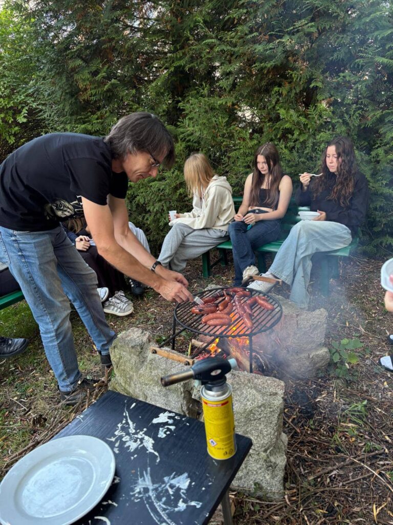 Grupa wychowanków Bursy Szkolnej podczas wspólnego ogniska z okazji Dnia Chłopaka. Młodzież siedzi na ławkach w otoczeniu zieleni, a na grillu nad ogniskiem pieką się kiełbaski.