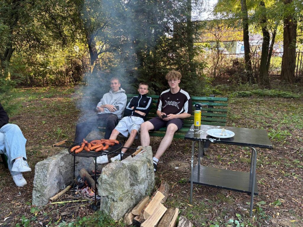 Grupa wychowanków Bursy Szkolnej podczas wspólnego ogniska z okazji Dnia Chłopaka. Młodzież siedzi na ławkach w otoczeniu zieleni, a na grillu nad ogniskiem pieką się kiełbaski.