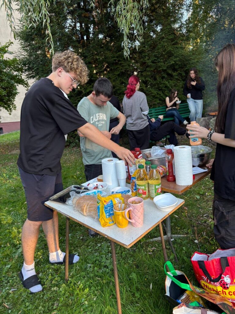 Grupa wychowanków Bursy Szkolnej podczas wspólnego ogniska z okazji Dnia Chłopaka. Młodzież siedzi na ławkach w otoczeniu zieleni, a na grillu nad ogniskiem pieką się kiełbaski.