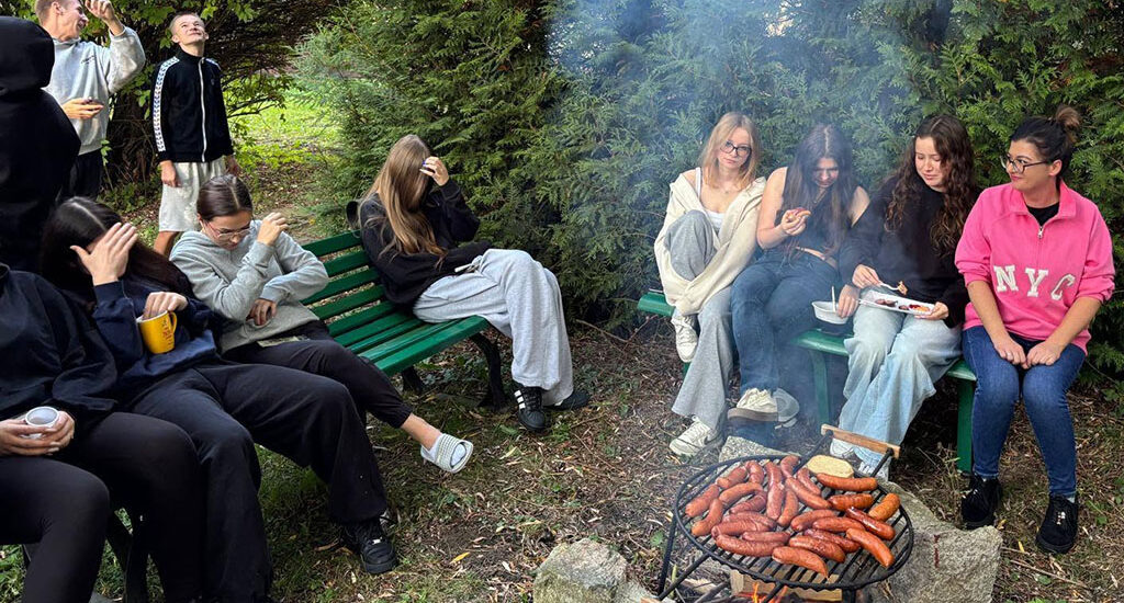 Grupa wychowanków Bursy Szkolnej podczas wspólnego ogniska z okazji Dnia Chłopaka. Młodzież siedzi na ławkach w otoczeniu zieleni, a na grillu nad ogniskiem pieką się kiełbaski.