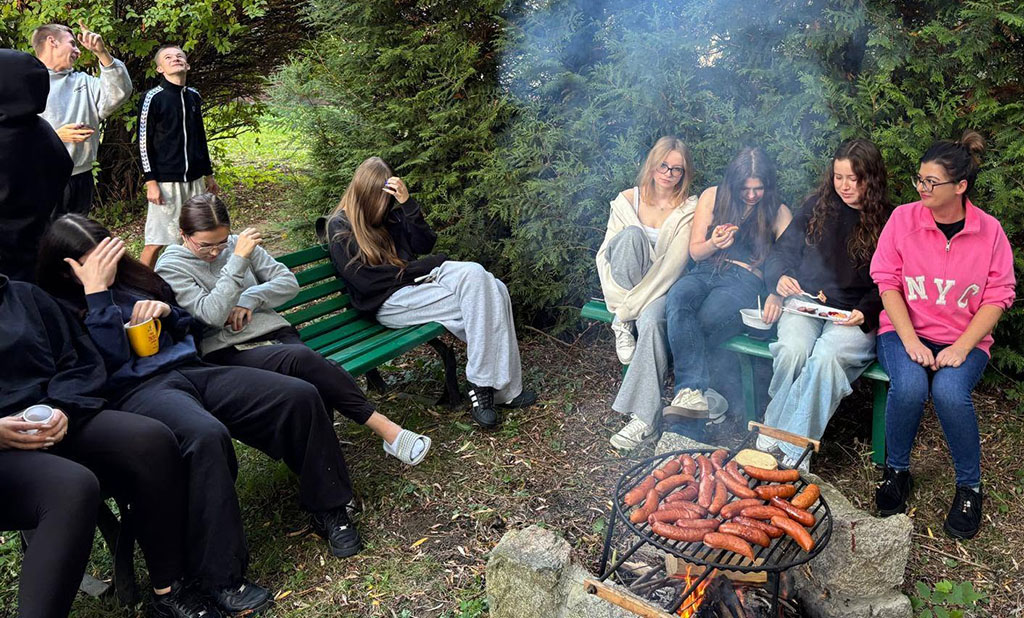 Grupa wychowanków Bursy Szkolnej podczas wspólnego ogniska z okazji Dnia Chłopaka. Młodzież siedzi na ławkach w otoczeniu zieleni, a na grillu nad ogniskiem pieką się kiełbaski.